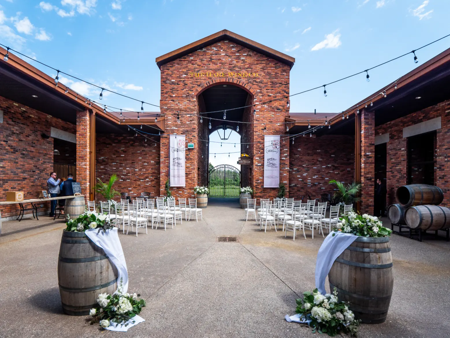 Rows of white chairs facing the brick archway, barrels and florals along the aisle