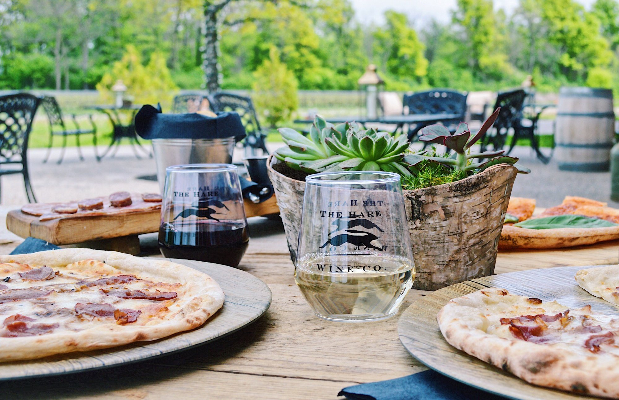 A table on the patio with wood-fired pizza and Hare glasses, vines behind