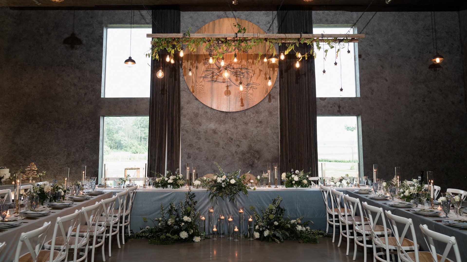Long tables set for a wedding reception, a wooden disc and hanging bulbs behind the head table