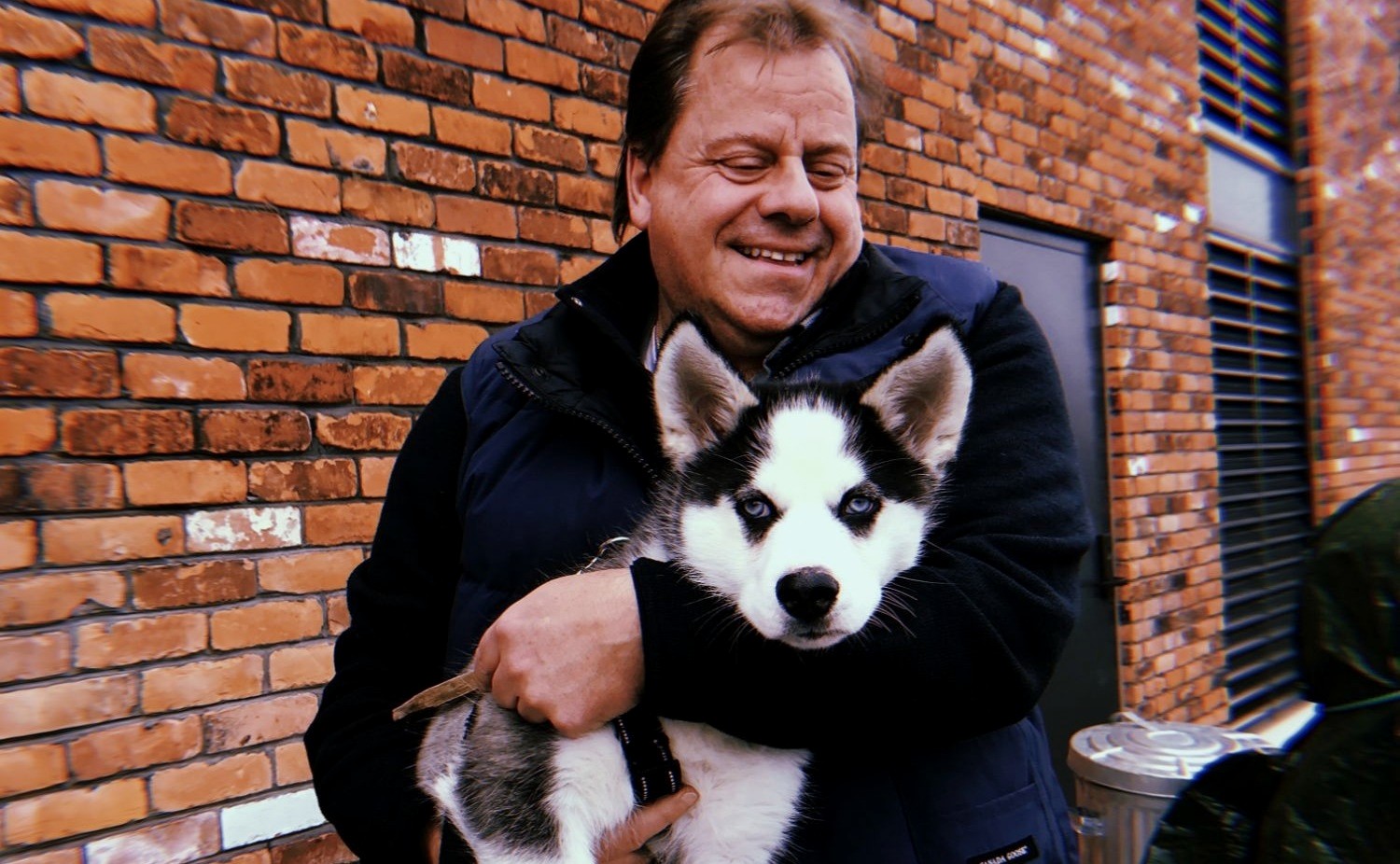 John Hare holding a husky puppy against the winery’s brick wall