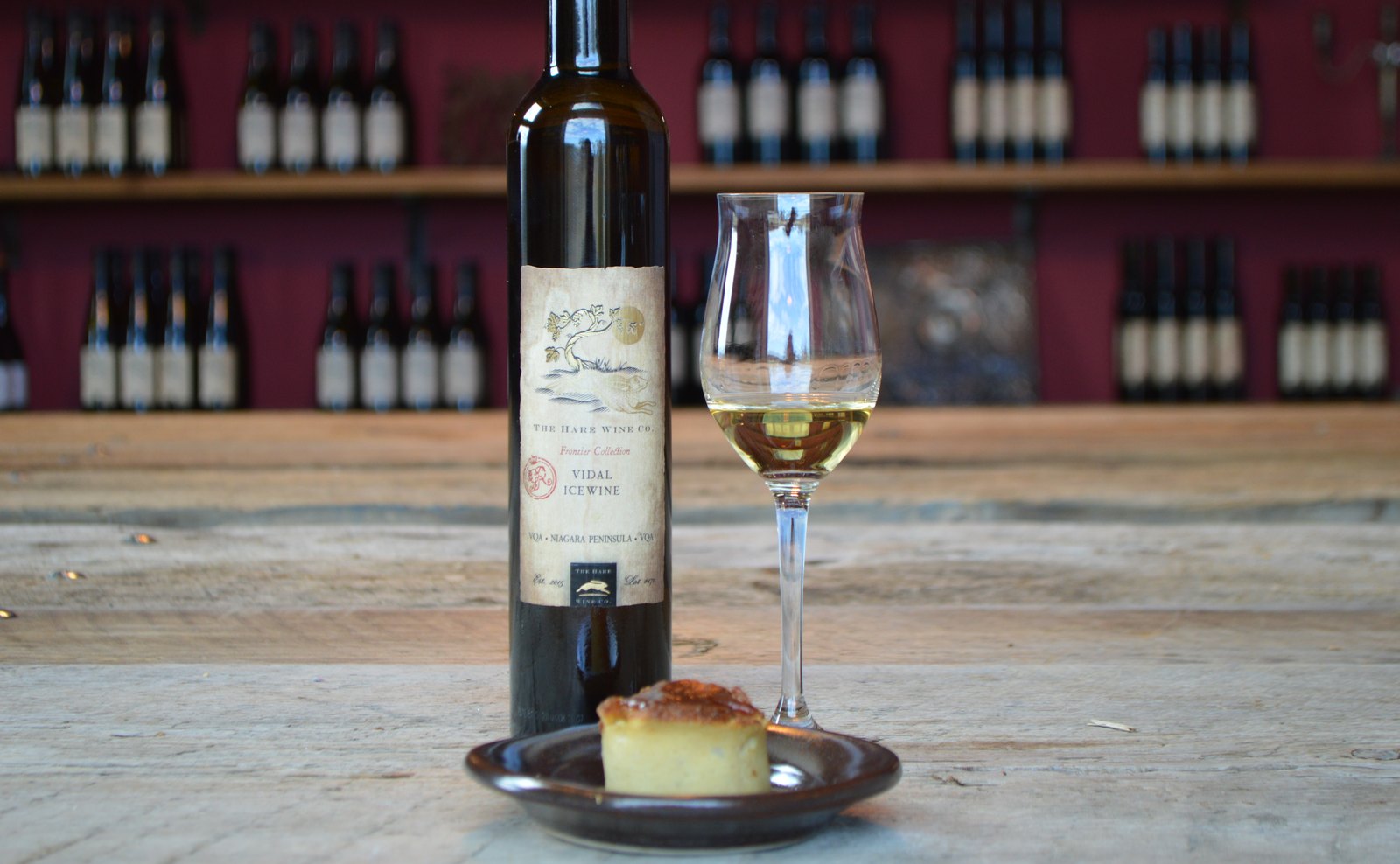 A glass of icewine and its bottle beside a cheese plate, wine racks behind