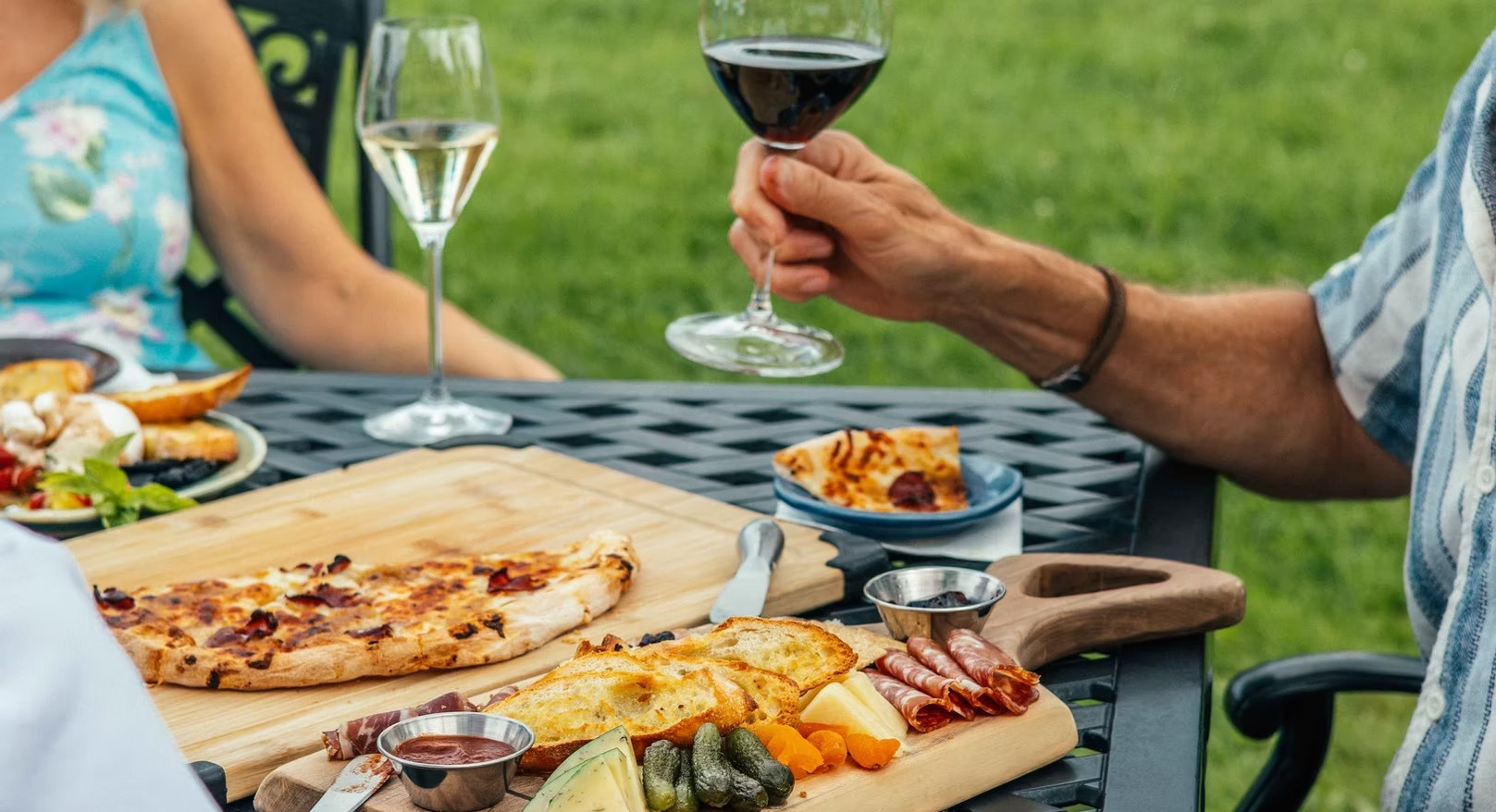 Hands with wine over an outdoor table of wood-fired pizza and a shared board