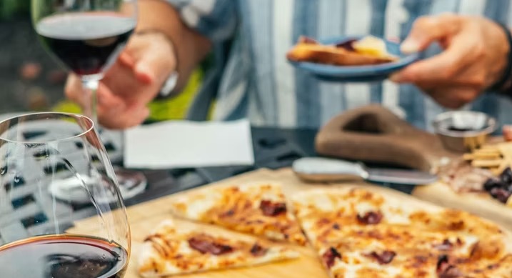 A glass of red raised over a table of wood-fired pizza